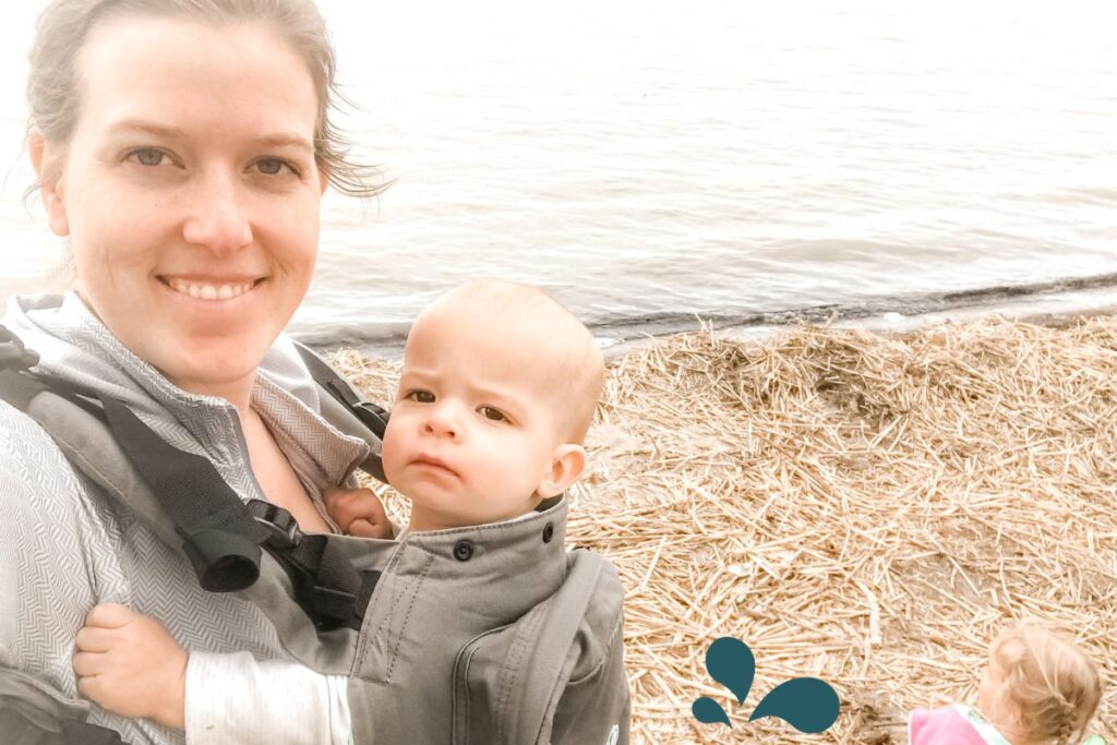 A contented mother smiles for a selfie by the shore, with her baby snug in a carrier, gazing curiously at the camera. In the background, another young child plays by the water’s edge, against a tranquil backdrop of calm waters and a straw-covered shoreline, encapsulating a peaceful family outing.