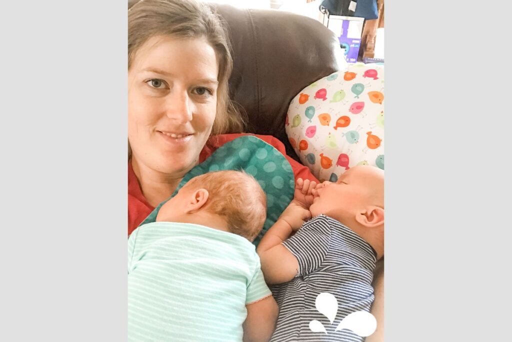 A smiling mother takes a selfie while comfortably reclining on a couch with her sleeping twin babies nestled against her, one in a green outfit and the other in blue stripes. The backdrop features a playful, fish-patterned nursing pillow, creating a cozy and tender family scene.