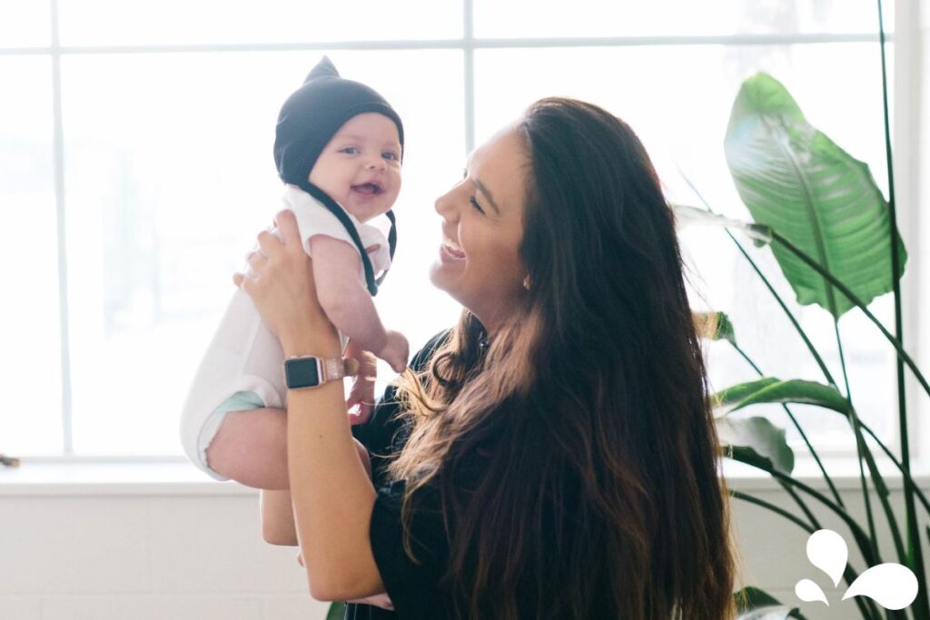 A radiant mother, with long dark hair and a smartwatch, holding her smiling baby who is adorably dressed in a white outfit with a black beanie. They are indoors, near a window with daylight streaming in, enhancing the warm, joyful atmosphere. A lush green plant adds a touch of nature to the scene.
