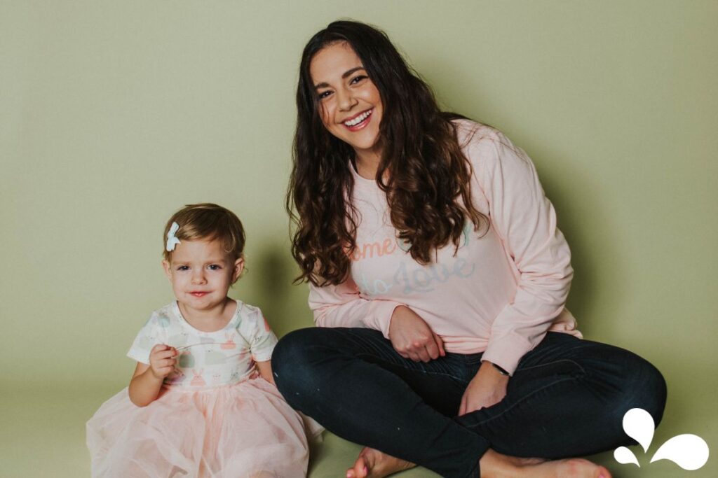 A smiling mother with long, wavy hair wearing a pink 'home is love' sweatshirt sits beside her young daughter, who is clad in a white and pink floral top with a fluffy pink skirt. The child has a sweet expression and is holding a small object, her hair adorned with a white bow. They pose against a soft green background that radiates warmth and familial love.