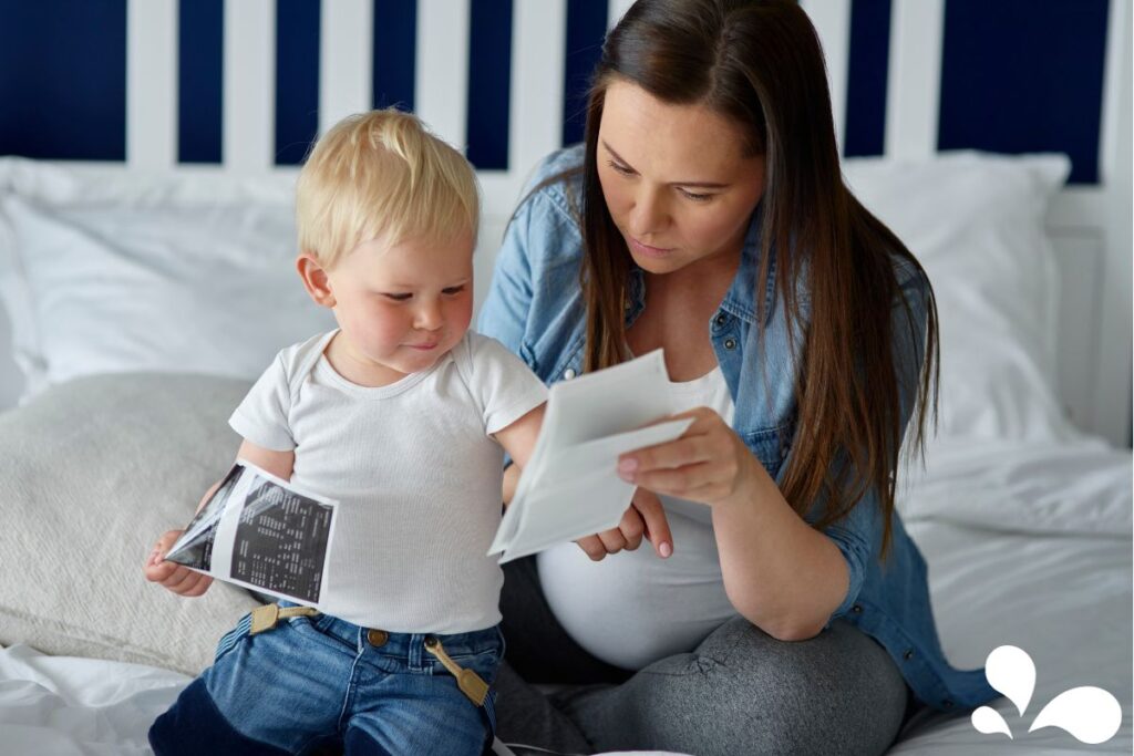 A young toddler, with an inquisitive expression, holds a sonogram image while sitting beside his pregnant mother on a bed. The mother, wearing a casual denim shirt and comfortable grey maternity leggings, is looking down at a piece of paper, perhaps another sonogram or related document. They are in a well-lit room with a white bed and dark blue headboard, creating a serene and intimate family moment.