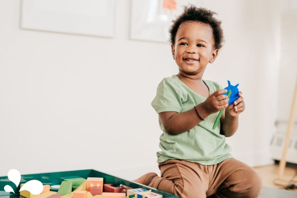 A cheerful toddler with a bright smile and curly hair sits on the floor holding a blue toy fish. Dressed in a light green shirt and brown pants, the child is surrounded by colorful blocks and shapes, indicating a playful learning environment. The child's delighted expression and the array of toys suggest a moment of pure, simple joy in the midst of playtime.