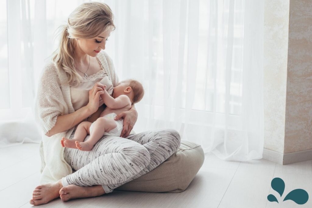 A tranquil image of a mother sitting on a pillow on the floor by a window, tenderly breastfeeding her baby. The soft natural light casts a gentle glow on the peaceful scene, highlighting the intimate bond between mother and child.