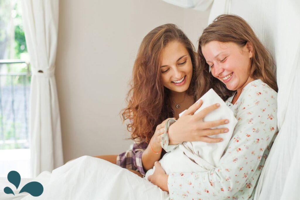 A friend smiles warmly as she visits a new mother who is cradling her swaddled baby, sharing a moment of joy and support.