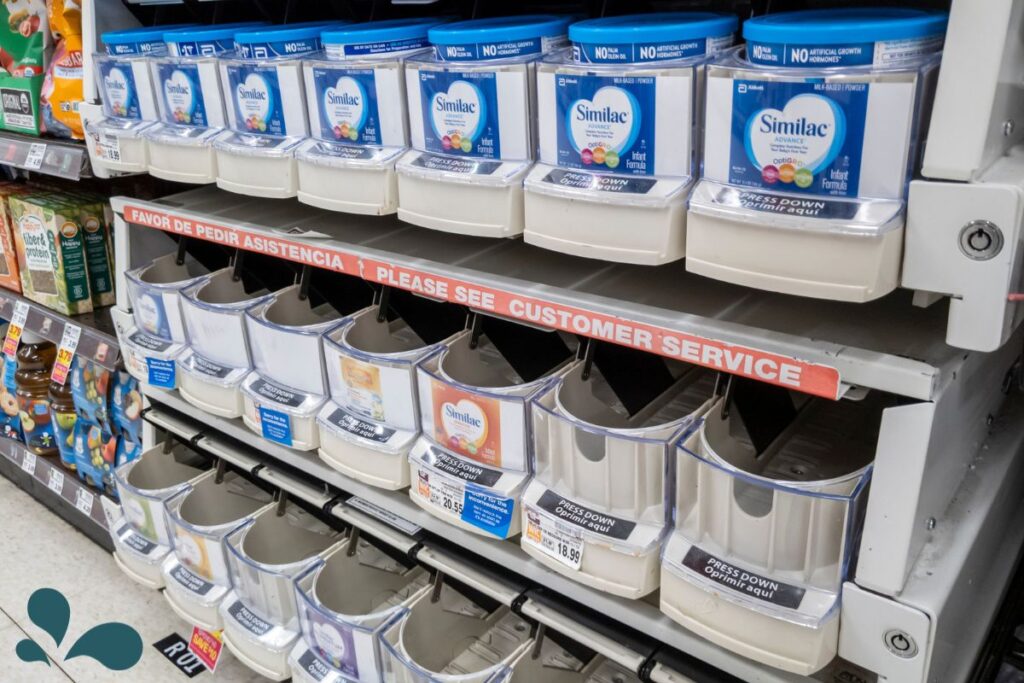 Shelves in a store with low stock of various brands of baby formula, indicating a shortage, with a sign requesting customers to see customer service.
