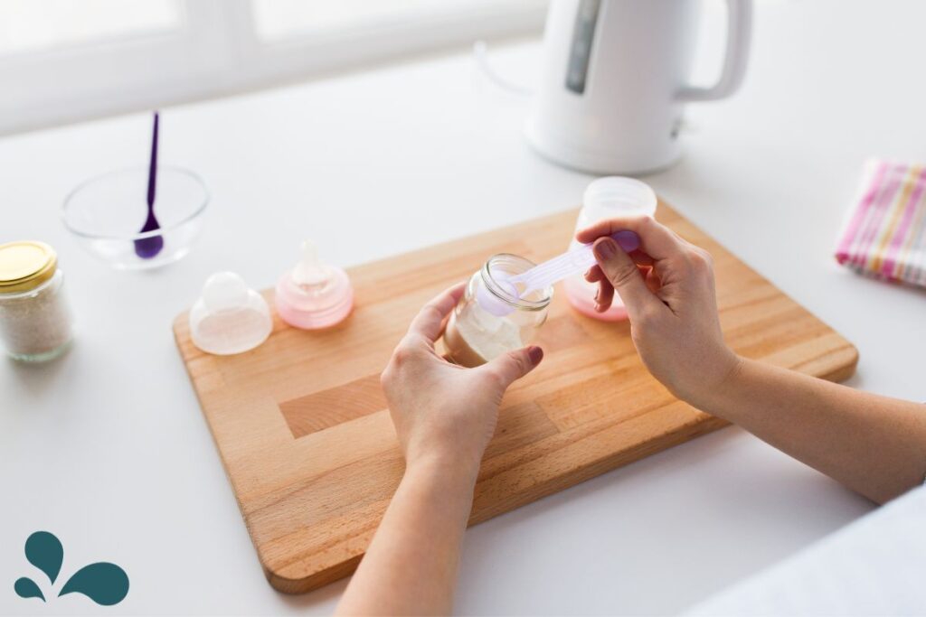 A woman preparing infant formula by pouring powder from a scoop into a baby bottle on a wooden cutting board near a kitchen window.