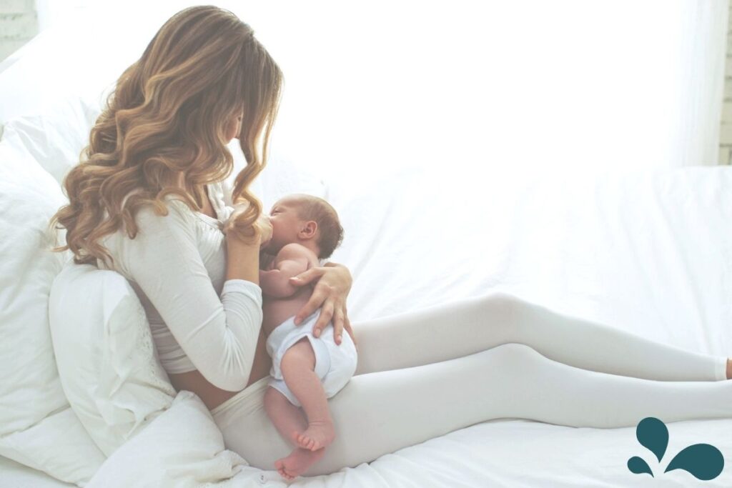Mom sitting up on a white linen-covered bed, breastfeeding newborn in a diaper with natural light coming through the window.