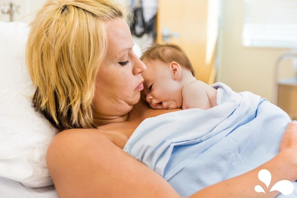 Mom and baby, skin-to-skin, in the hospital during the golden hour. Mom is laid back in the hospital bed with baby on her chest and covered with blue blankets.