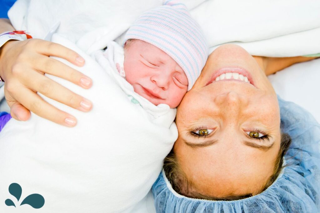 A new mother in a hospital gown smiles brightly as she lies back, cradling her newborn baby who is wrapped in a swaddle and wearing a striped hat.