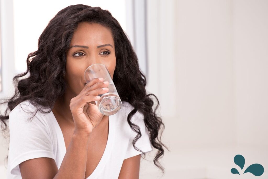 A woman with long curly hair, wearing a white t-shirt, drinking a glass of water, symbolizing the importance of staying hydrated.