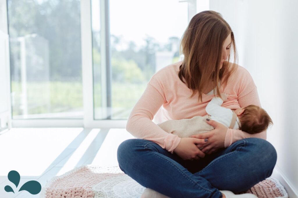 A woman seated by a bright window, wearing a peach-colored sweater and blue jeans, breastfeeding her infant, who is wrapped in a white blanket.