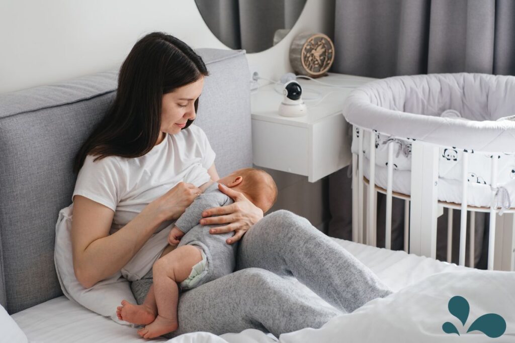 A mother sitting on a bed, breastfeeding her baby, with a bassinet in the background. She is wearing a white t-shirt and gray sweatpants, looking affectionately at her infant.