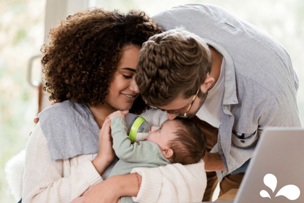 A tender family moment with a mother bottle-feeding her baby, embraced by the father who is leaning in to kiss the baby's head. The parents are smiling gently, radiating warmth and affection in a softly lit home setting.