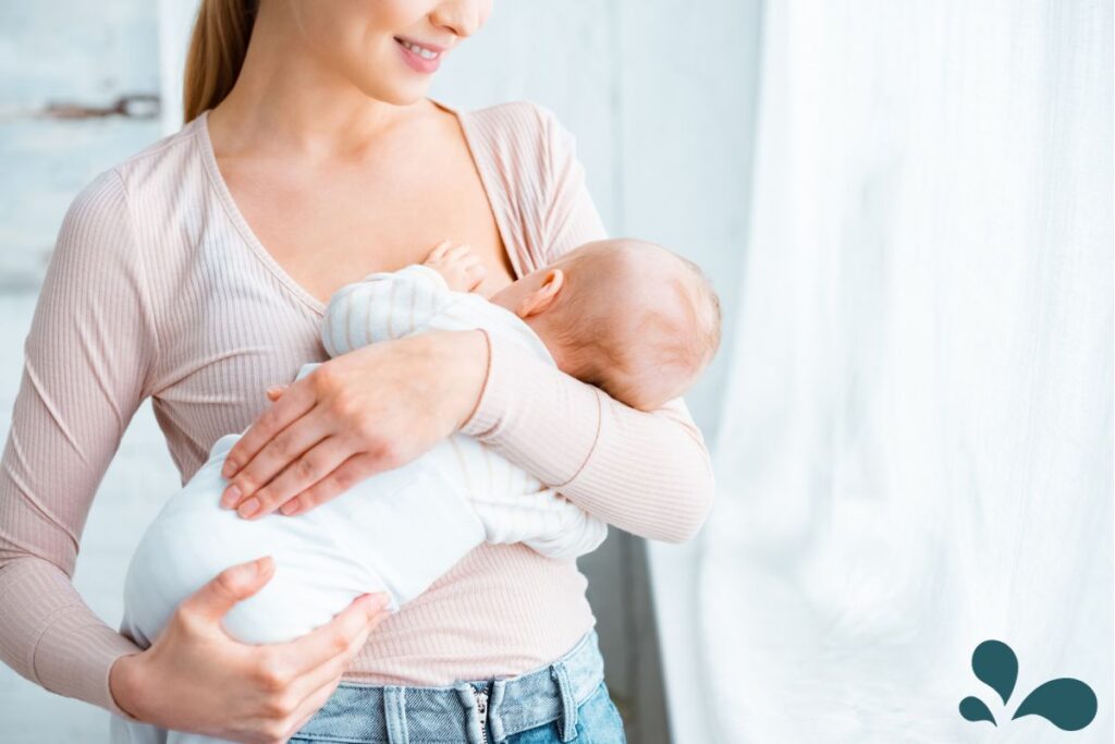 A smiling woman in a pink shirt lovingly holds and nurses her swaddled newborn baby in a gentle and nurturing embrace.