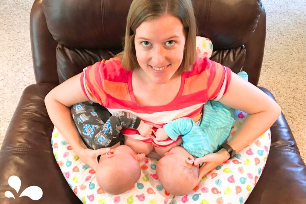 A smiling woman comfortably seated in a brown recliner breastfeeding twin babies, each swaddled in different colored blankets. The babies are resting on a colorful nursing pillow with a playful print, in a cozy, well-lit living room.