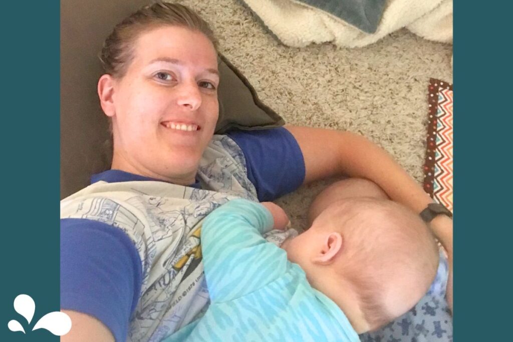 A mother lying on her side takes a selfie while breastfeeding her baby, who is also lying down facing her. Both are relaxed and comfortable on a carpeted floor, with a pillow behind the mother and a colorful throw cushion nearby.