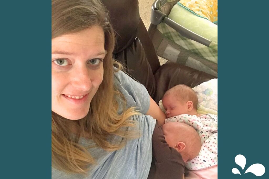 A smiling mother takes a selfie while breastfeeding one of her twins, with the second twin cradled in her other arm. Both babies are asleep and comfortably snuggled against her, in a relaxed home environment with a baby playpen in the background.