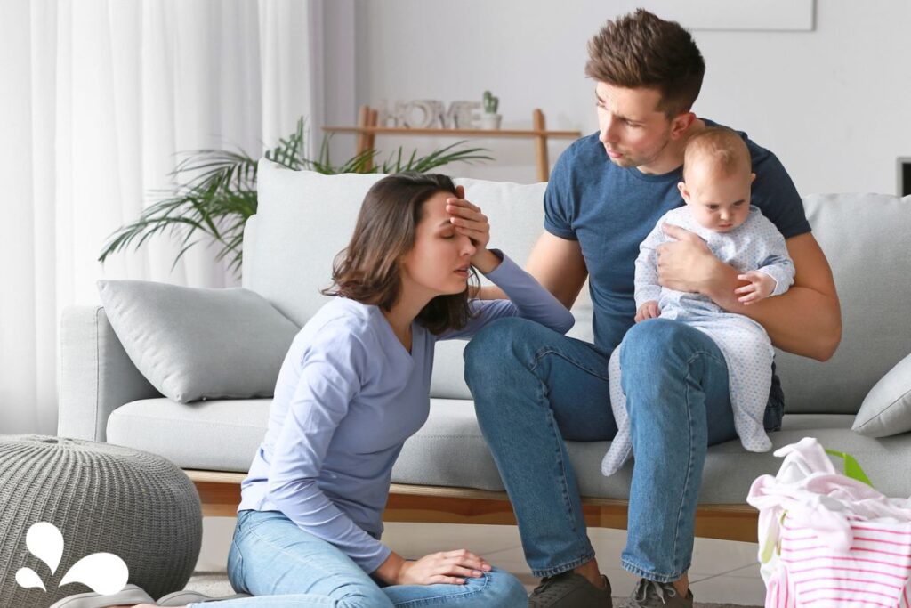A concerned father seated on a sofa, holding a baby, turns towards the mother who appears stressed and is sitting on the floor with her hand on her forehead. The room is well-lit with a comfortable home setting and a basket of laundry is visible in the foreground.