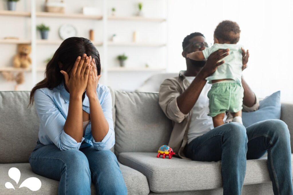 A distressed mother covers her face with her hands, sitting on a sofa, while the father, sitting beside her, affectionately holds their baby up to his face. The living room is cozy with shelves and a child's toy on the sofa, suggesting a family environment.