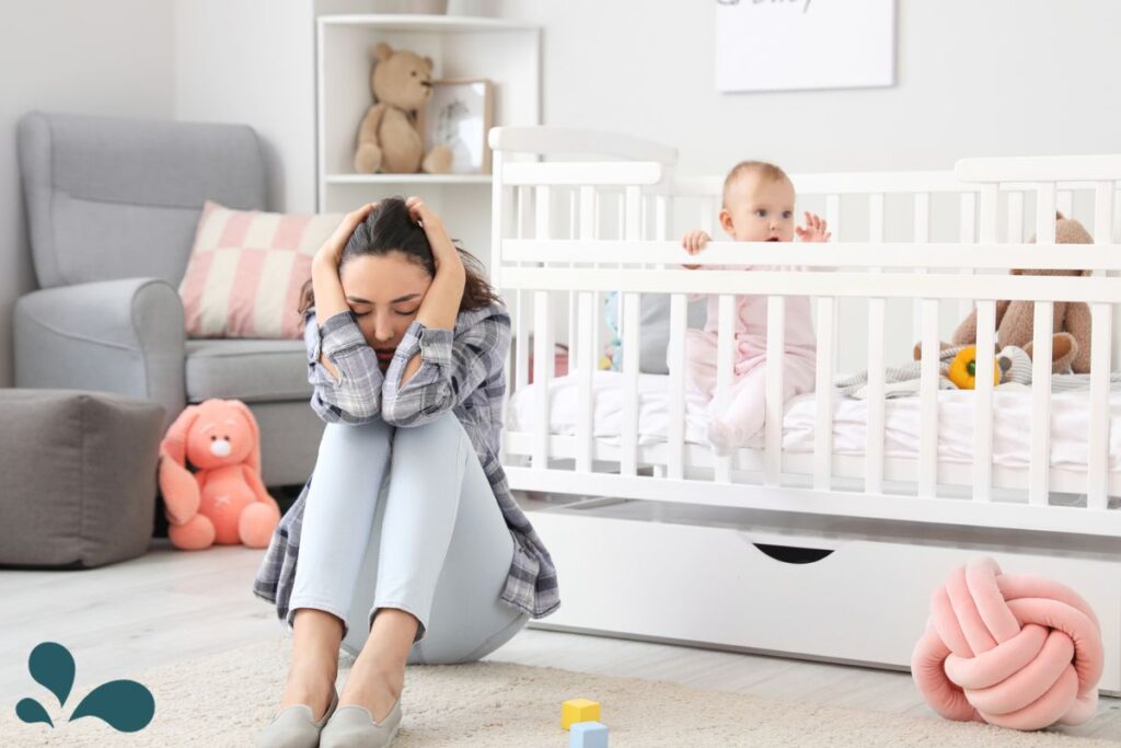 A distressed mother sits on the floor with her head in her hands, while in the background, a baby sits up in a white crib, observing. The room is softly lit and decorated with a plush bear, a knot cushion, and children’s toys, creating a comforting nursery environment.