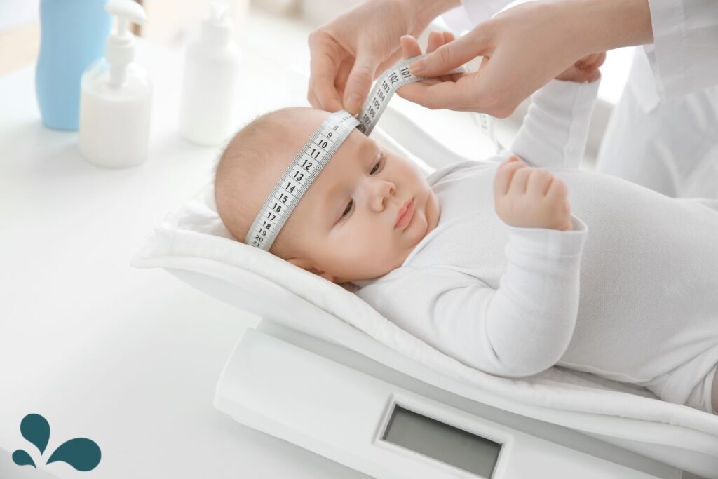 A healthcare professional measures the head circumference of a calm baby lying down, using a measuring tape. The infant is dressed in white, resting on a scale with a digital display, in a clinical setting with bottles of care products in the background.