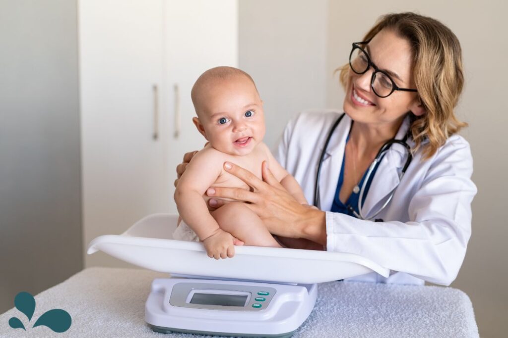 A happy baby with blue eyes and a slight smile is being held on a pediatric scale by a smiling female doctor with glasses and a stethoscope around her neck. The scale's display is visible in the foreground, set against a clean, clinical background.