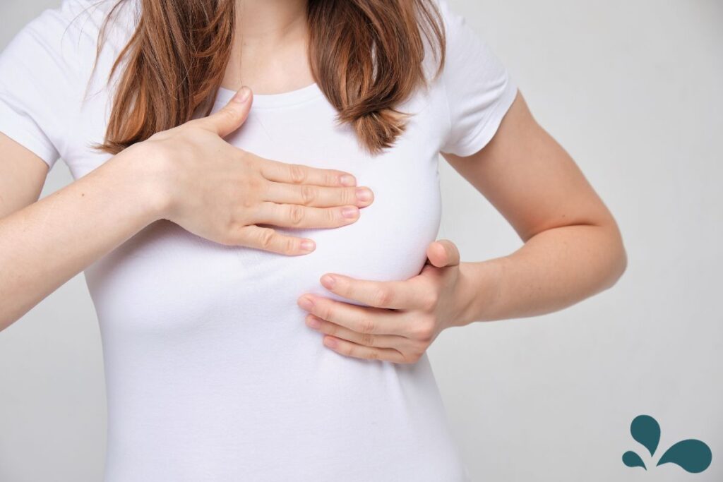 A close-up of a woman in a white shirt cradling her chest, with hands placed gently to suggest discomfort or breast pain.