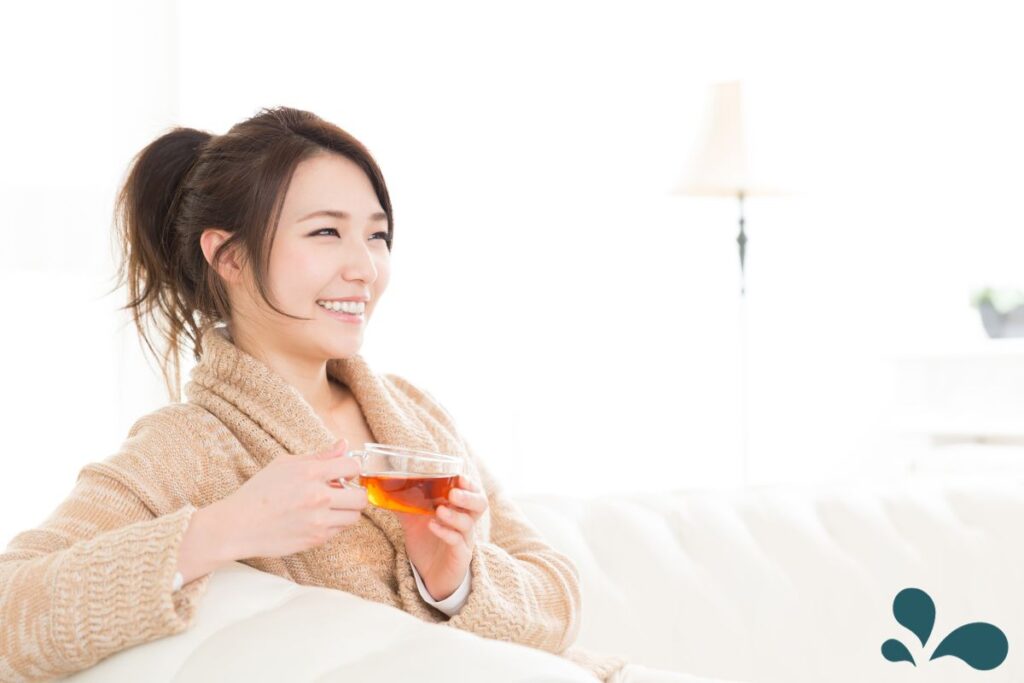 A cheerful woman in a cozy robe holding a glass cup of tea, with a warm smile and relaxed posture.