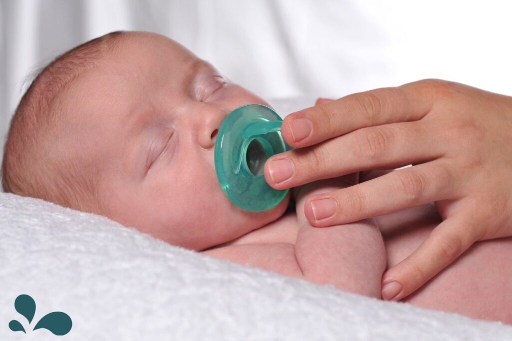 A newborn baby peacefully sleeping with a green pacifier in their mouth, held gently by an adult's hand.