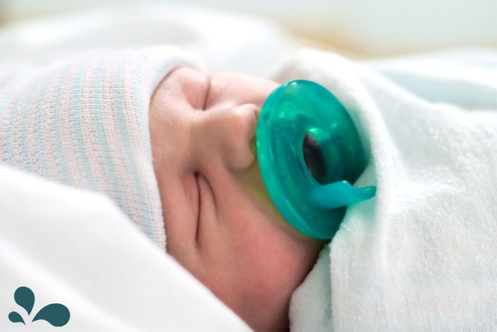 A swaddled newborn baby with a teal pacifier, resting calmly on a soft white surface.