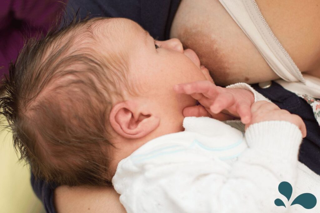 A close-up view of a newborn with a shallow latch during breastfeeding, the baby's hand gently touching the mother's breast.