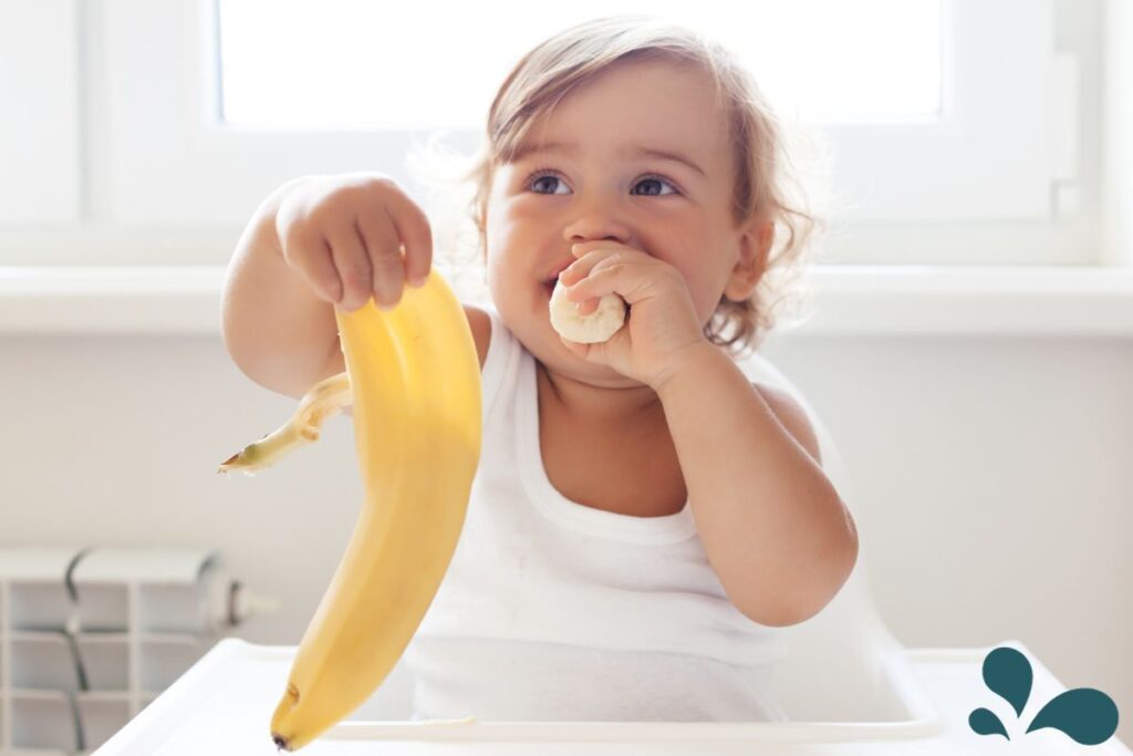 A smiling baby in a high chair reaching for a banana, experiencing the delight of tasting new foods.