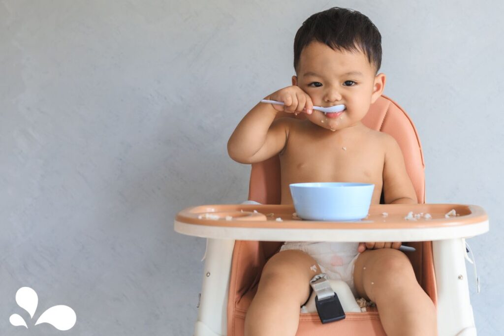 A cheerful baby using a spoon to feed themselves from a blue bowl, enjoying the messy milestone of self-feeding.